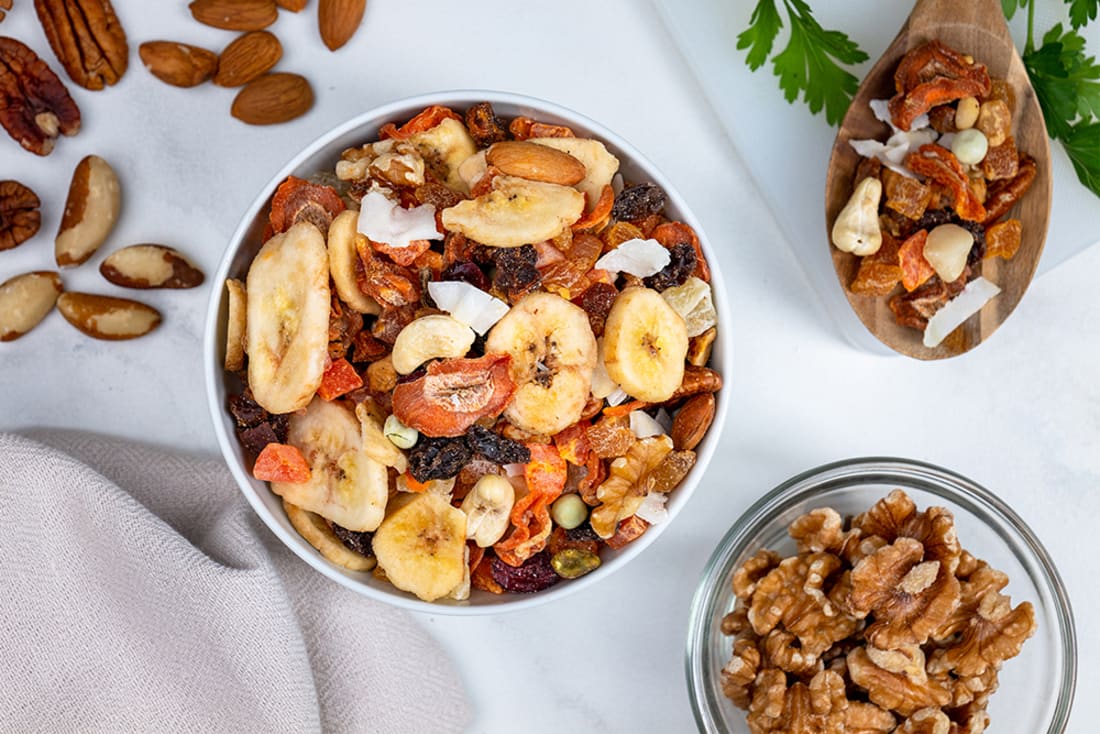 Trail mix with nuts, dried fruits, and bananas in a bowl on a white surface.