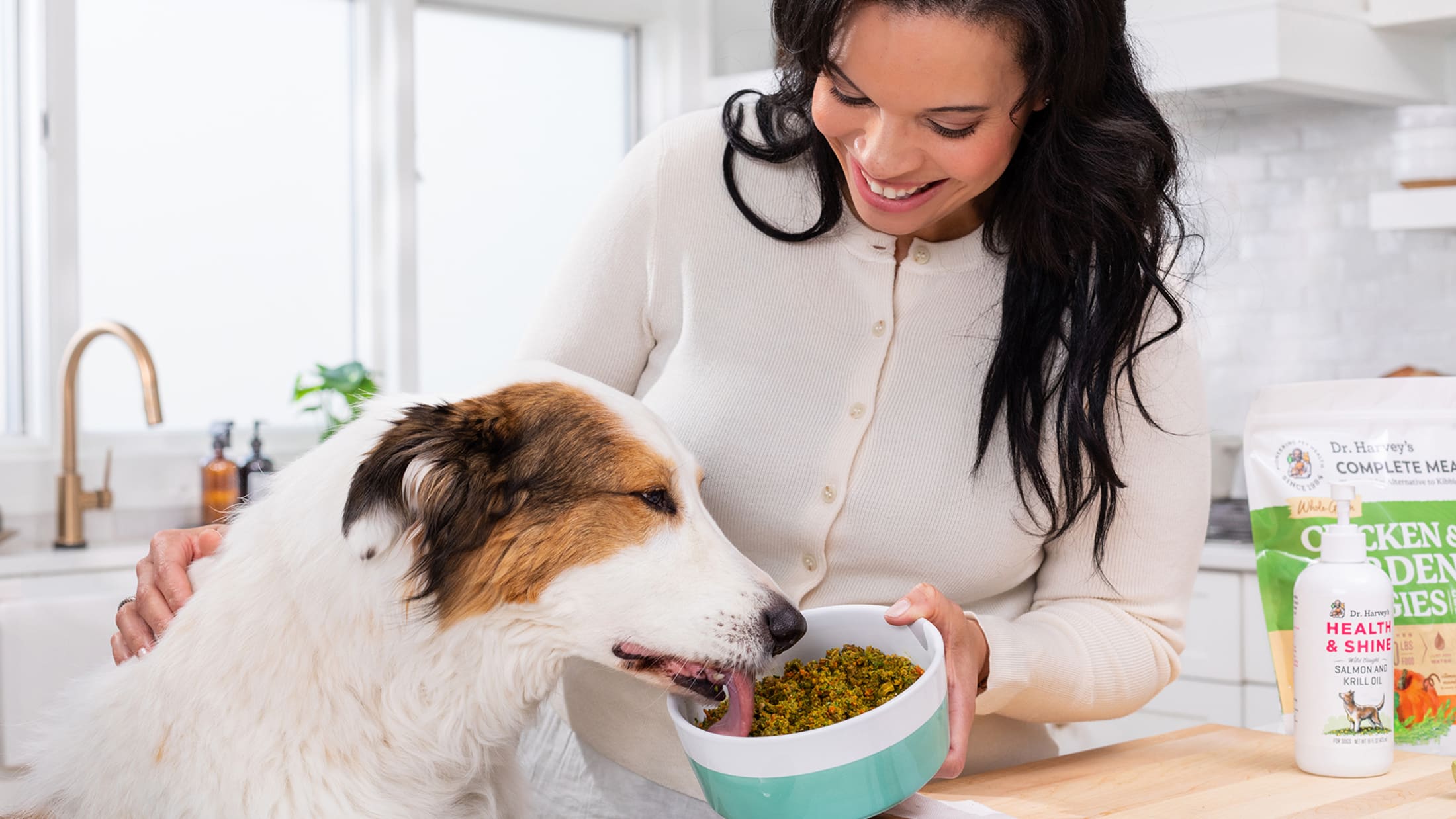 Woman feeding a dog from a bowl in a kitchen with pet food packaging visible